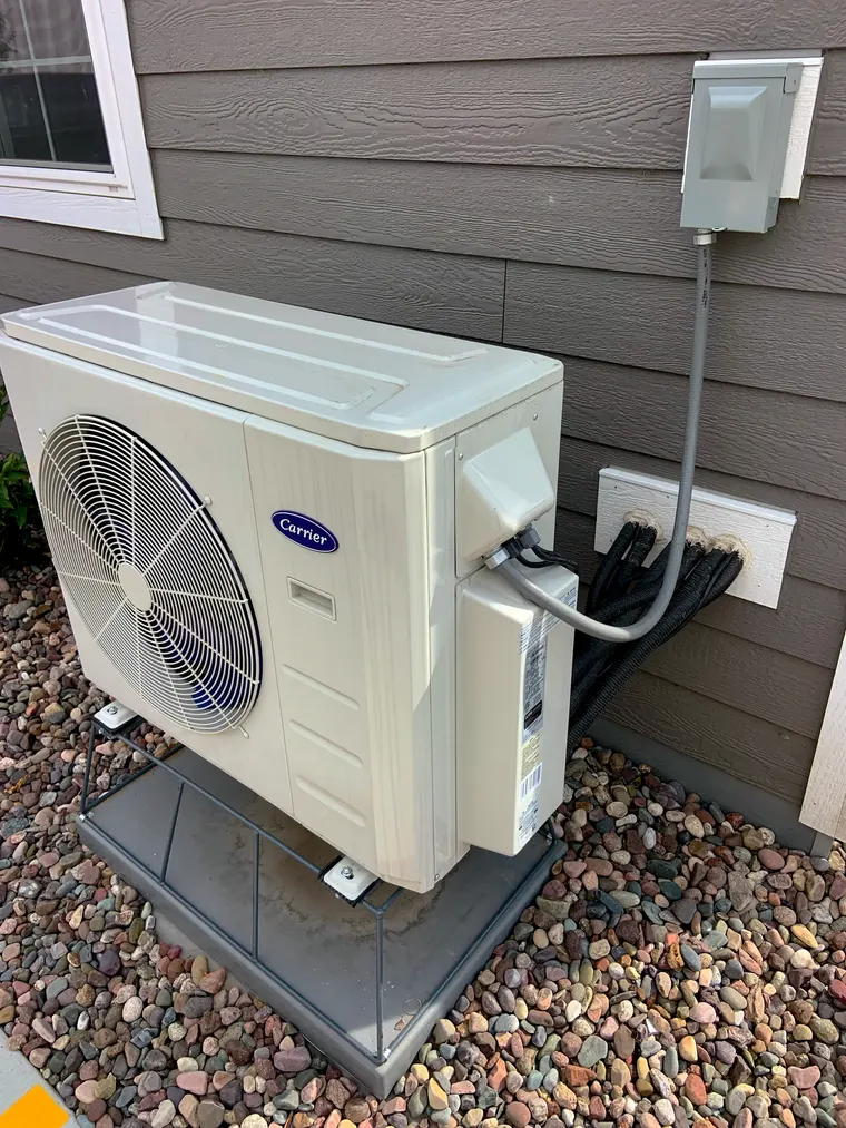 An air source heat pump outdoor unit standing on a concrete pad beside a house wall, with its fan grille facing outward.